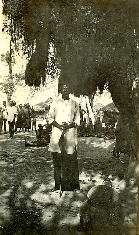 Chief Mkanya standing beneath a tree at his village, people gathered behind