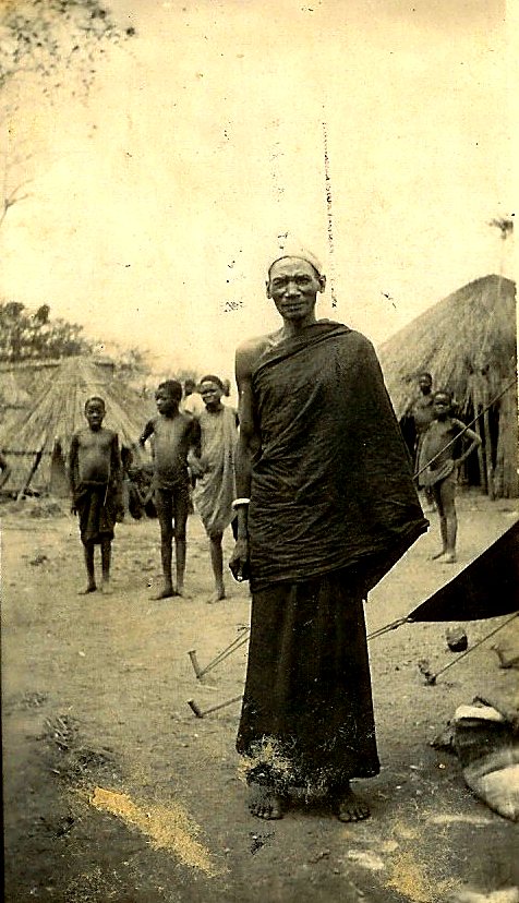 Chief Vumbe standing in his village, children and huts behind him